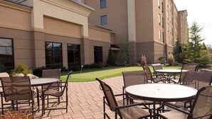 An outdoor patio area with round tables and chairs, a well-maintained lawn, and a building with a beige facade and large windows. - Hampton Inn & Suites Vineland (Vineland)