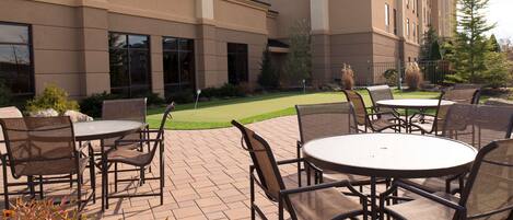 An outdoor patio area with round tables and chairs, a well-maintained lawn, and a building with a beige facade and large windows.