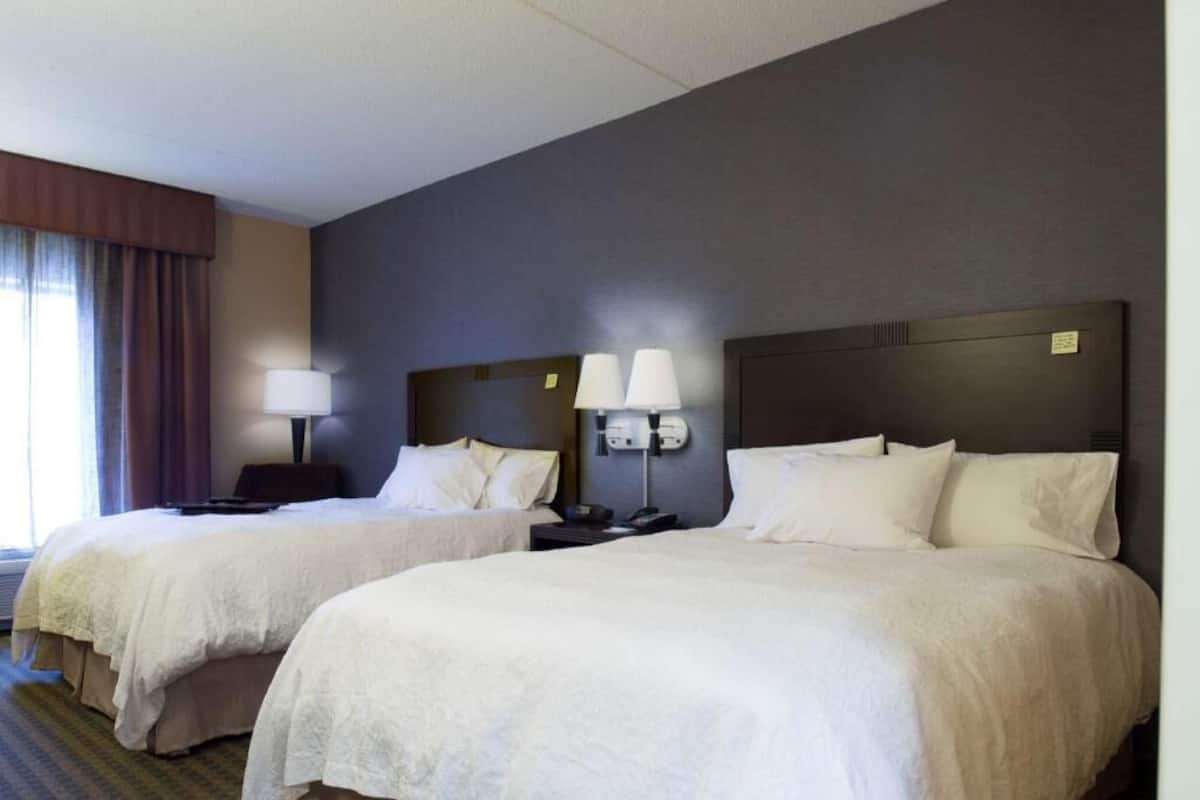 A hotel room with two beds, a desk, a lamp, and a window with curtains.