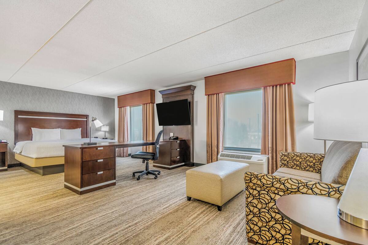 A hotel room with a bed, desk, chair, ottoman, and a TV.