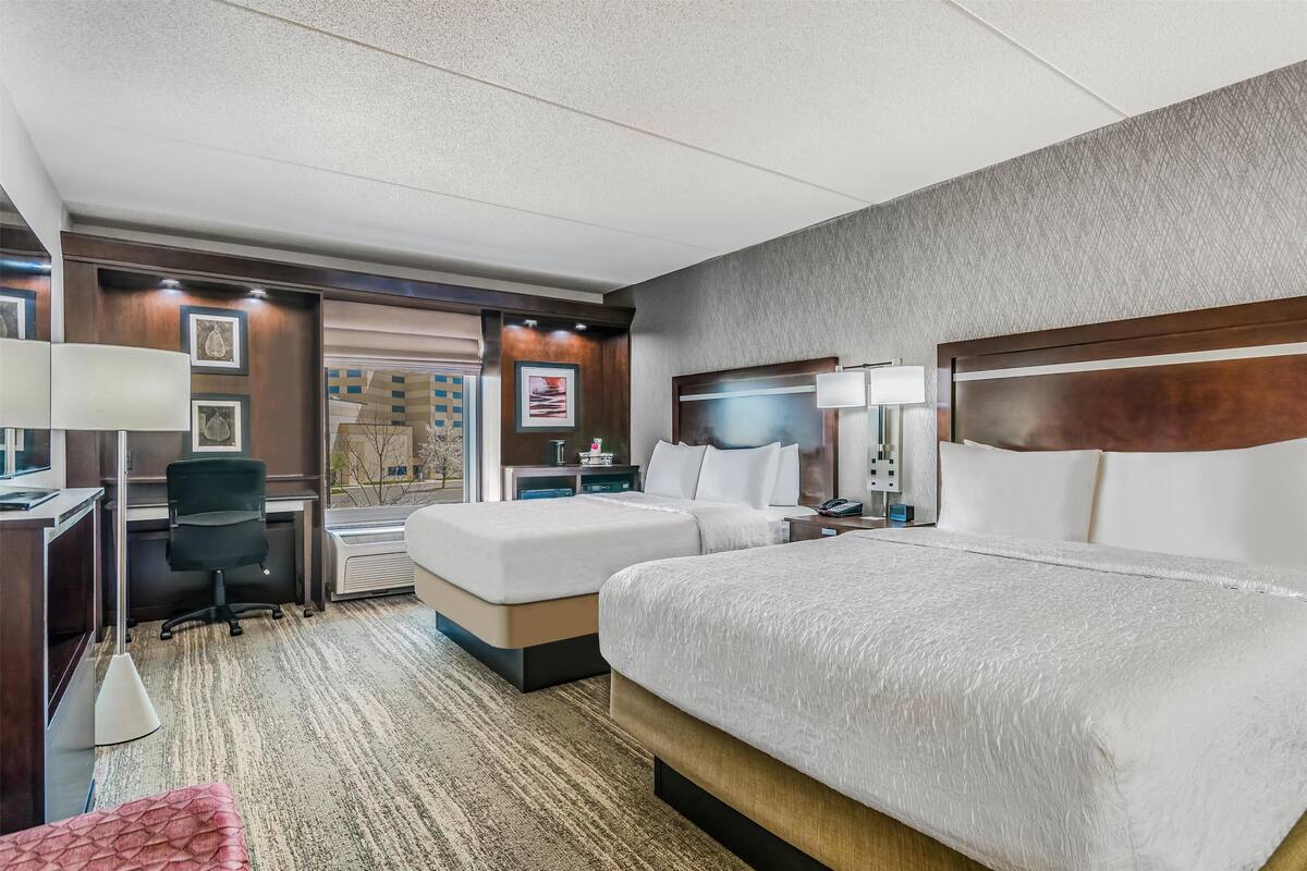 A hotel room with two beds, a desk, a chair, and a large window.