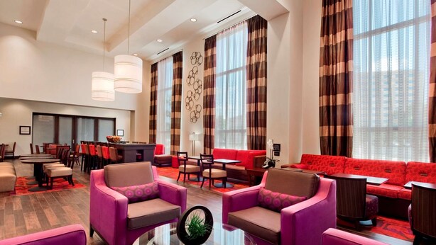 Un hall d’hôtel moderne avec un canapé rouge, des fauteuils violets, un coin repas et de grandes fenêtres ornées de rideaux à rayures.