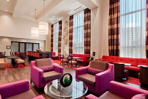 A modern hotel lobby with a red sofa, purple armchairs, a dining area, and large windows with striped curtains. - Hampton Inn & Suites Detroit / Airport - Romulus (Romulus)
