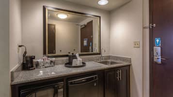 A hotel bathroom with a granite countertop, a coffee maker, a sink, a mirror, and a door with a keycard lock.
