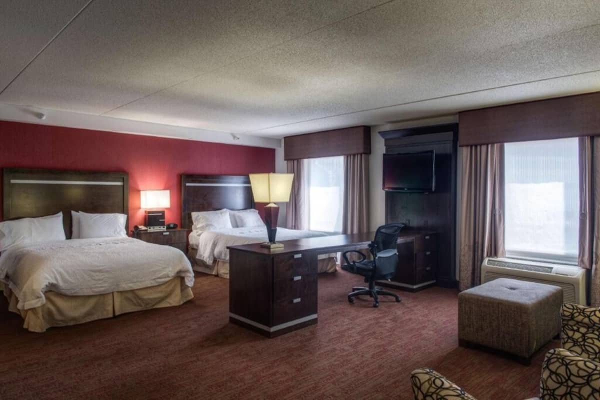 A hotel room with two beds, a desk, a chair, an ottoman, and a television.