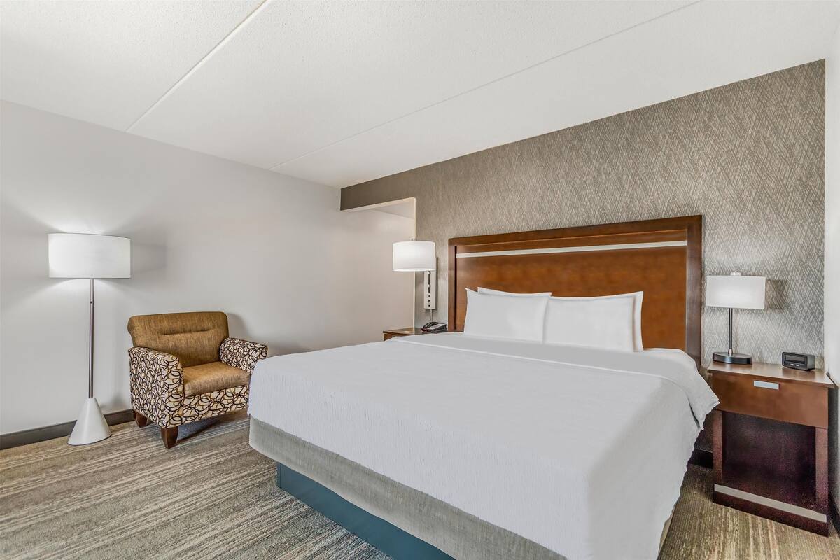 A hotel room with a large bed, a patterned armchair, a standing lamp, and a nightstand with a lamp.