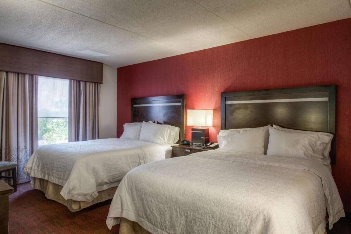A hotel room with two beds, a nightstand, a lamp, and a window with curtains.