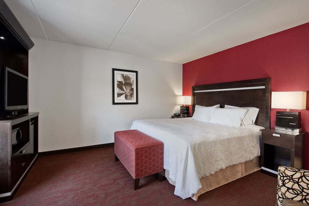 A hotel room with a bed, bedside tables, a TV, a red accent wall, and a framed picture on the wall.