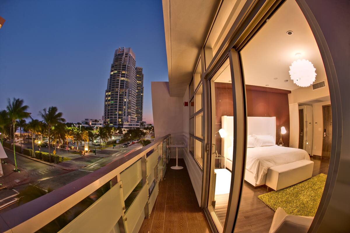A balcony with a city view, a bedroom with a bed and a nightstand, and a large window.