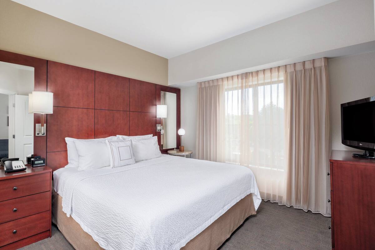 A hotel room with a large bed, a TV, a wooden headboard, a bedside table with a lamp, and a window with curtains.