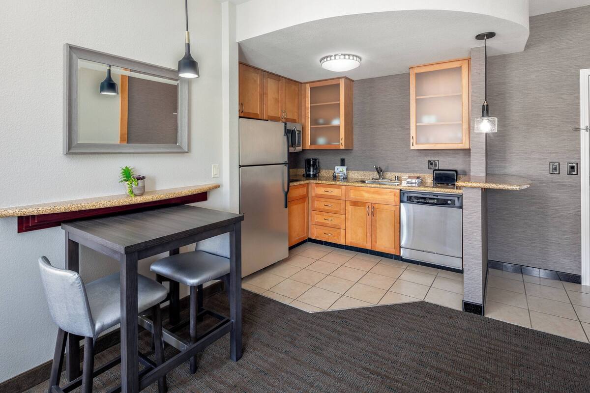 A compact kitchen with wooden cabinets, a stainless steel refrigerator, a dishwasher, and a dining area with a table and two chairs.