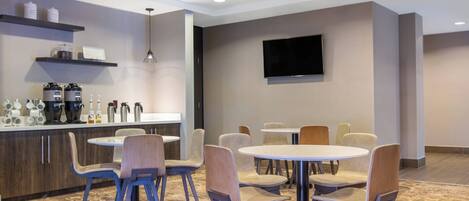 A modern hotel room with a kitchen area, a dining table with chairs, and a flat-screen TV mounted on the wall.
