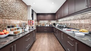 A modern kitchen with dark wood cabinets, granite countertops, and a central island with a coffee machine and food display. - Residence Inn Phoenix NW/Surprise (Surprise)