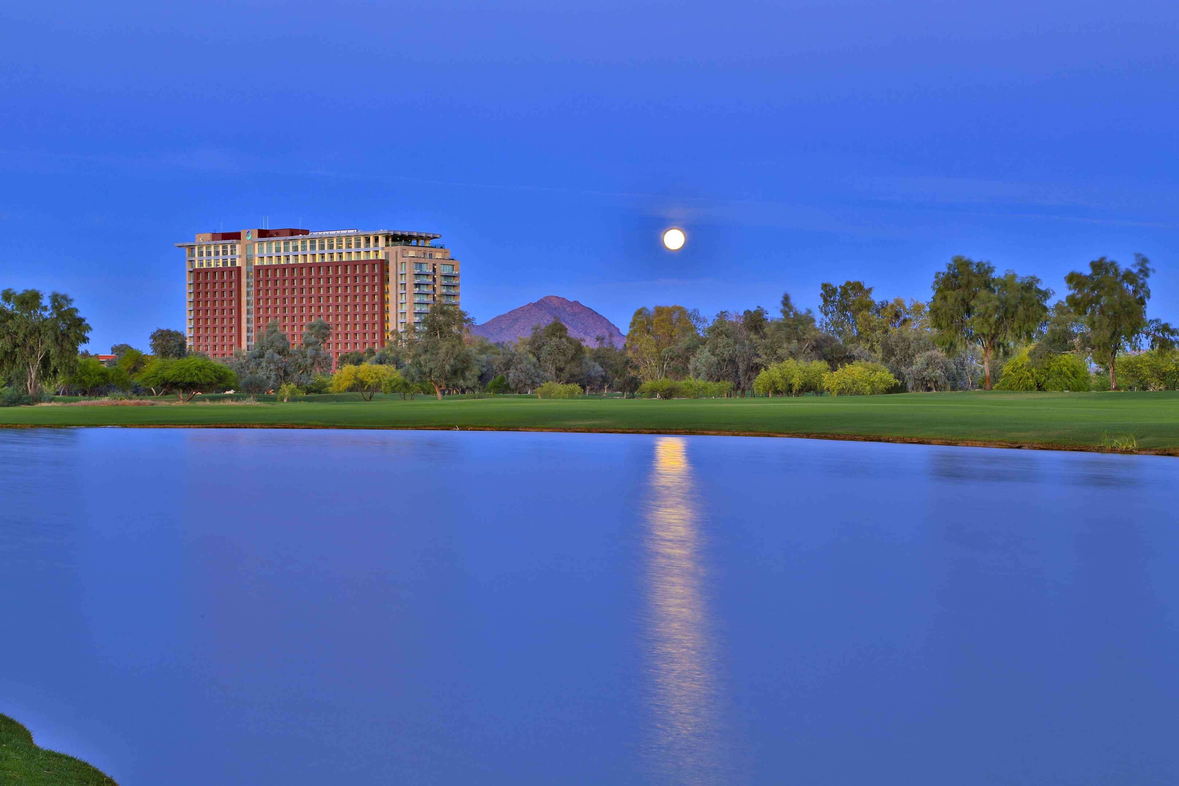 A large hotel with a mountain in the background, a body of water in the foreground, and trees around.