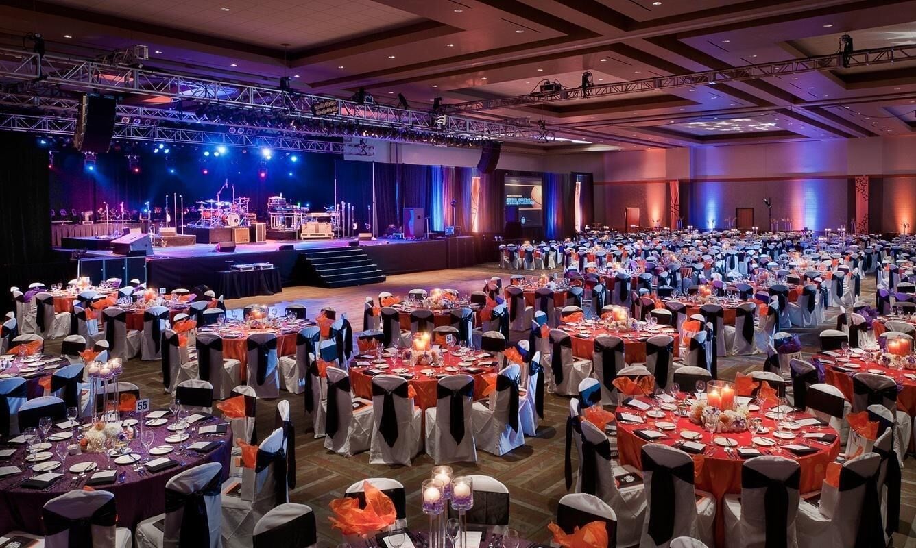A banquet hall with round tables set for a formal event, a stage with musical instruments, and a large audience seated.