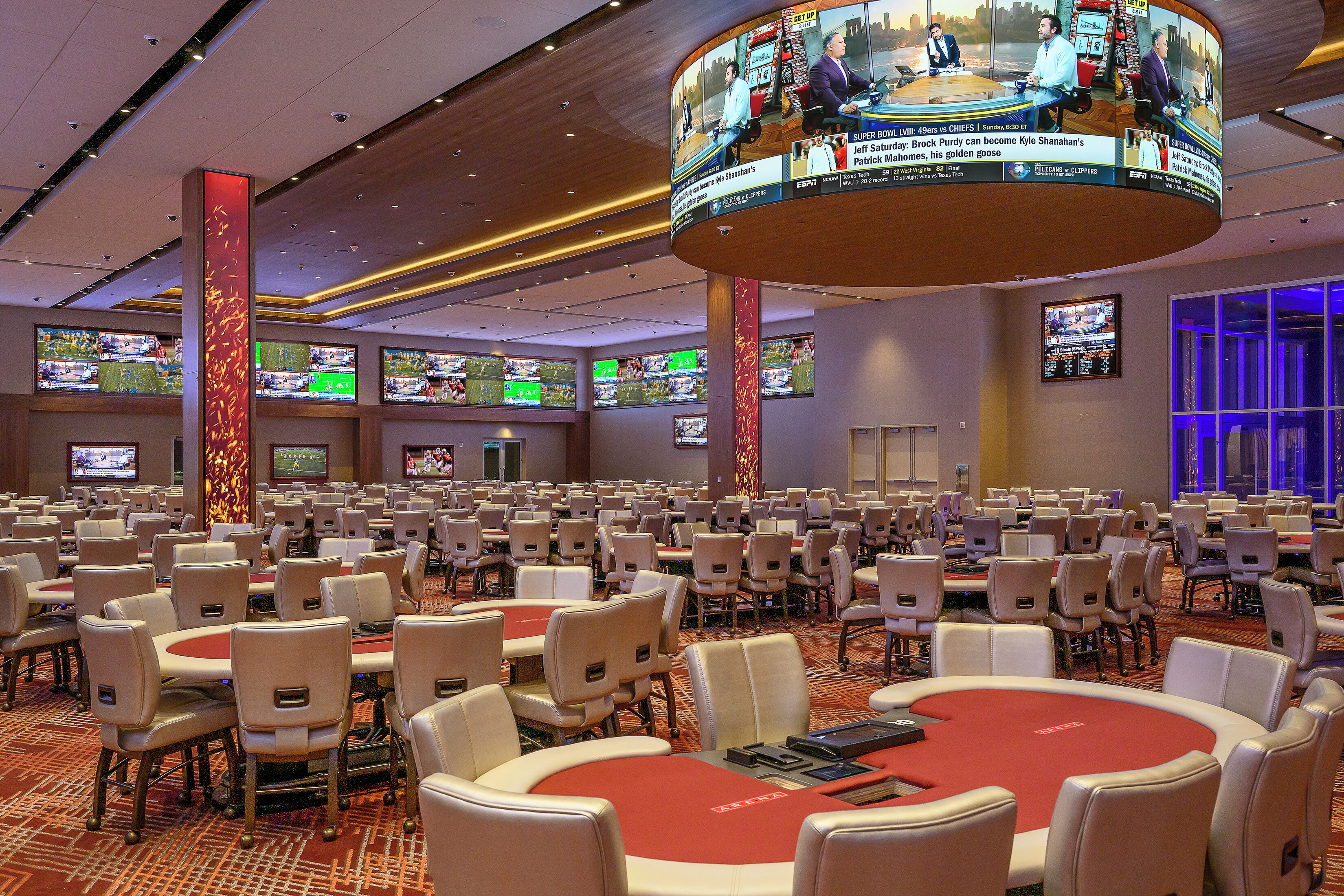 A large conference room with multiple tables and chairs, a central red poker table, and multiple screens displaying sports events.