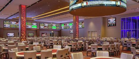 Een ruime congreszaal met meerdere tafels en stoelen, een rode poker tafel in het midden, en meerdere schermen waarop sportevenementen te zien zijn.