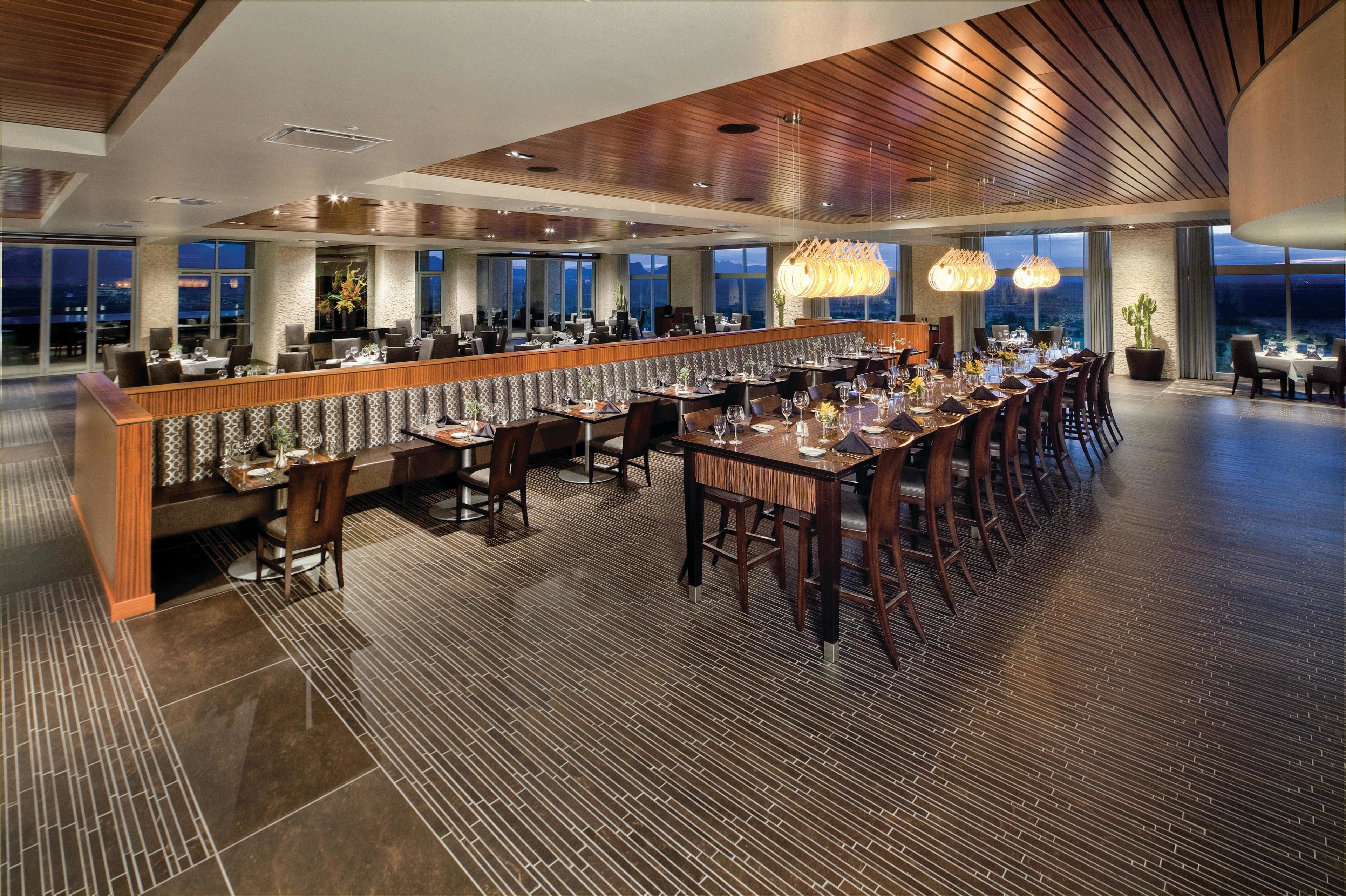 A modern restaurant with a long counter, multiple dining tables, and a high ceiling with wooden paneling.