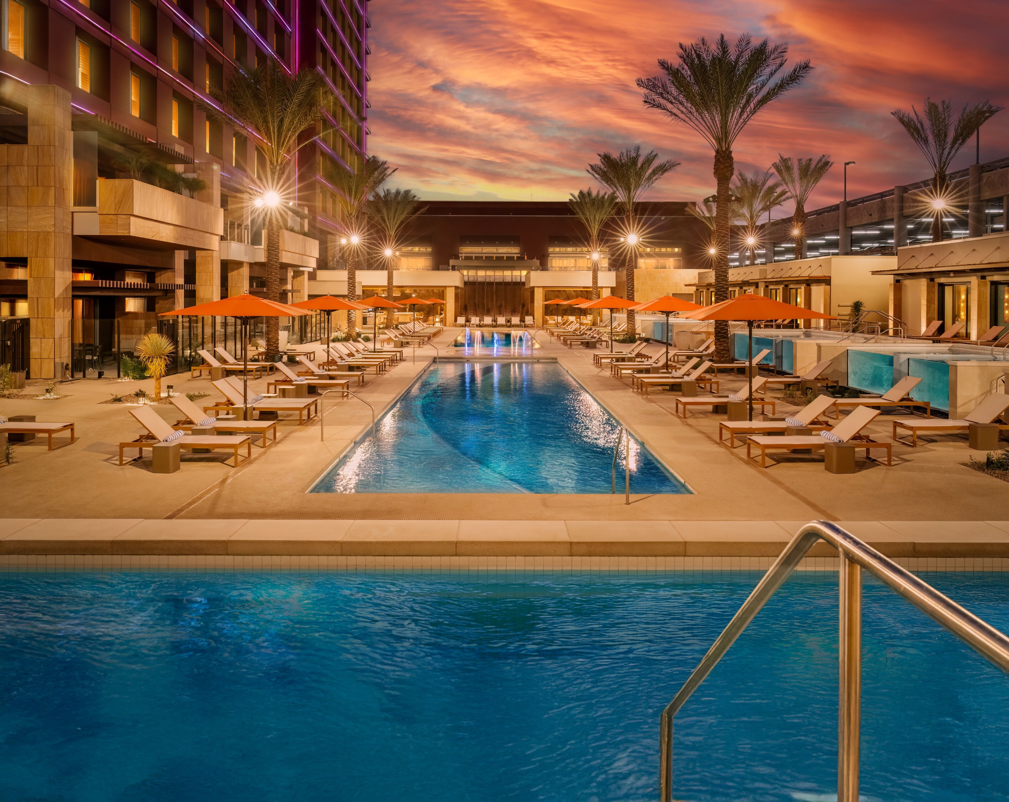 A swimming pool with lounge chairs and umbrellas, surrounded by palm trees and a hotel building with illuminated windows.