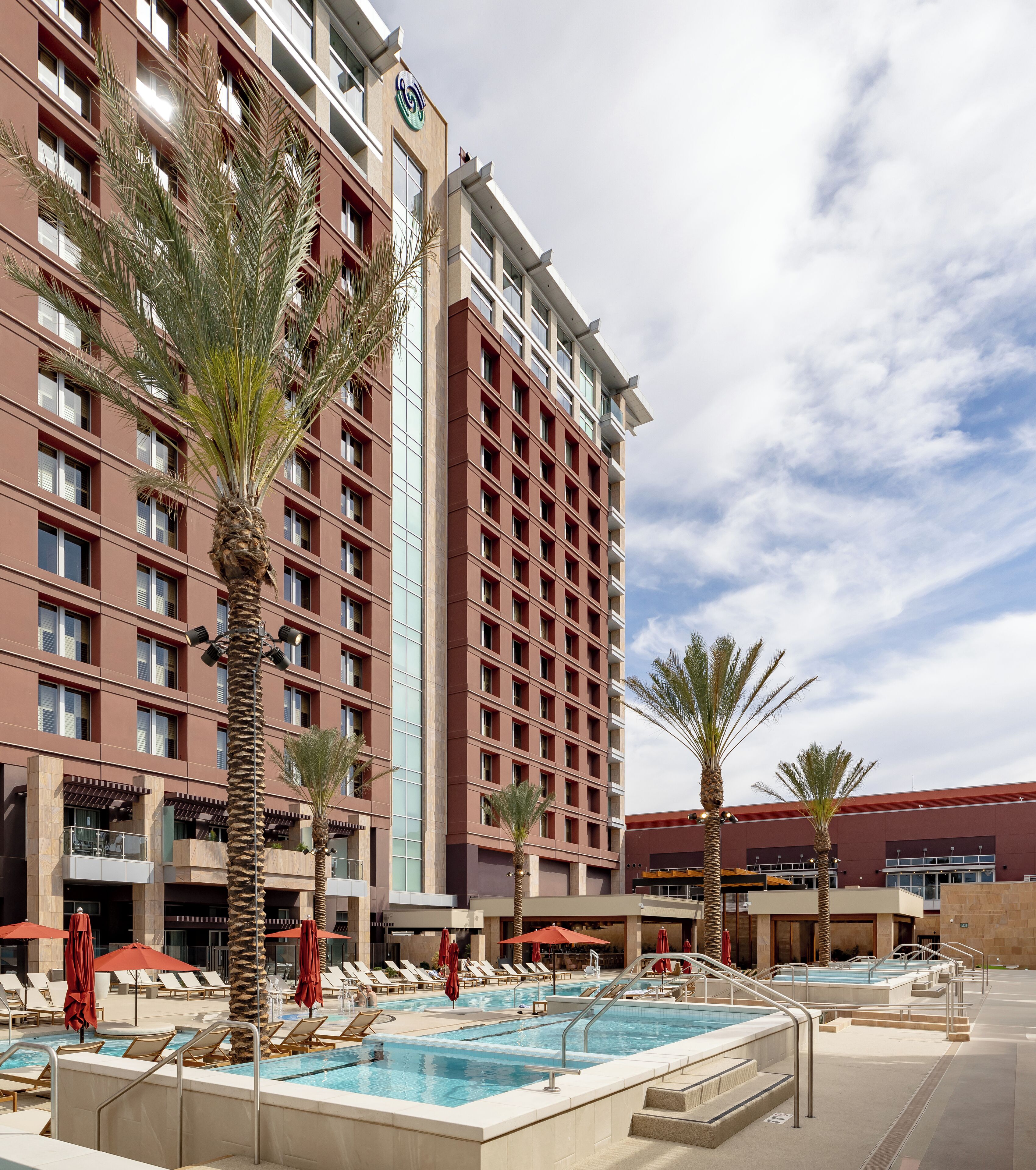 A hotel with multiple floors, a swimming pool area with sun loungers and umbrellas, and palm trees.