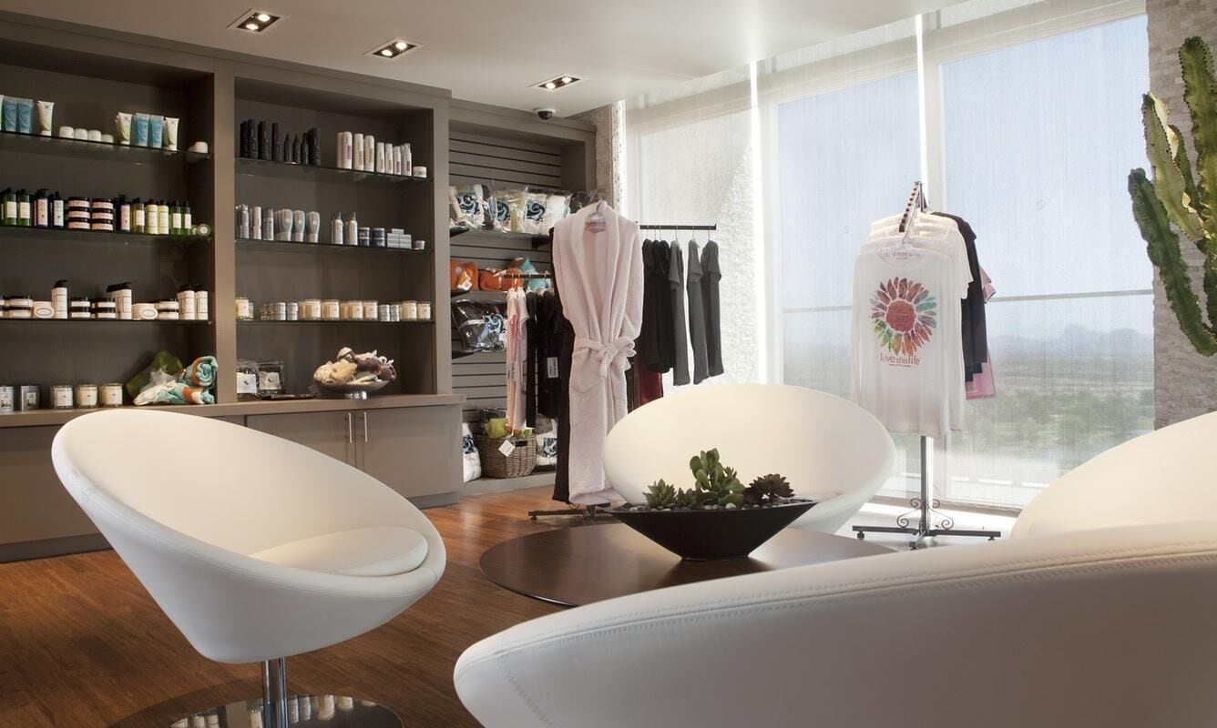 A modern spa room with a wooden floor, white chairs, a large window, and shelves with various spa products.