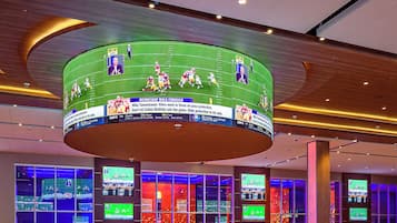 Un casino con múltiples mesas de blackjack, una gran pantalla central que muestra eventos deportivos y una decoración interior moderna.