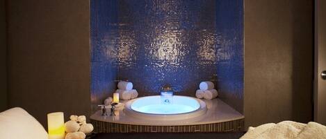 A modern spa room with a central hot tub, a dark tiled wall, and a black carpeted floor.