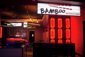 Un bar de fideos tenuemente iluminado, con un letrero de neón que dice 'Bamboo Noodle Bar' y una zona de estar con mesas y sillas.