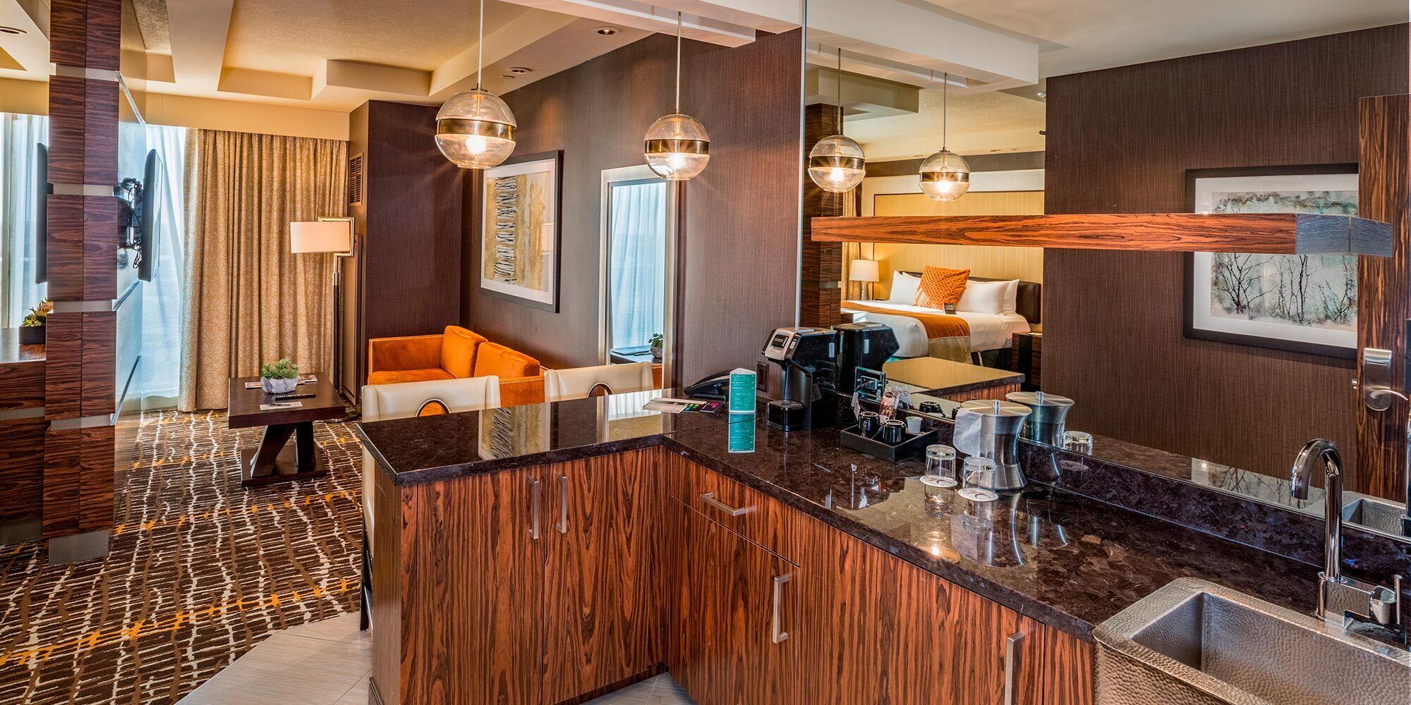 A modern hotel room with a wooden accent wall, a seating area with a sofa, a coffee station, and a large mirror.