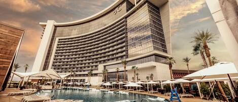 A hotel with a large pool, sun loungers, umbrellas, and palm trees.