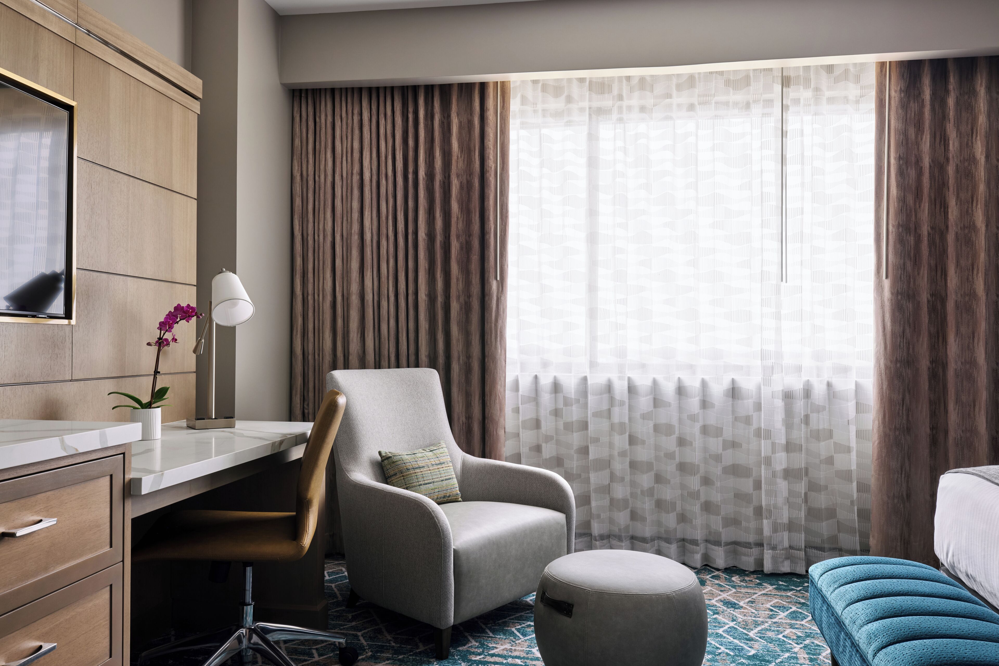 A hotel room with a desk, chair, ottoman, and a large window with curtains.