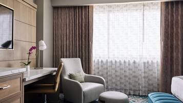 A hotel room with a desk, chair, ottoman, and a large window with curtains.