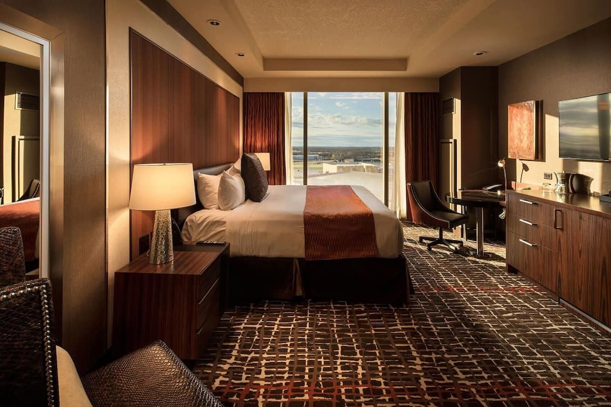 A hotel room with a large bed, a desk, a chair, a TV, and a view of the city.