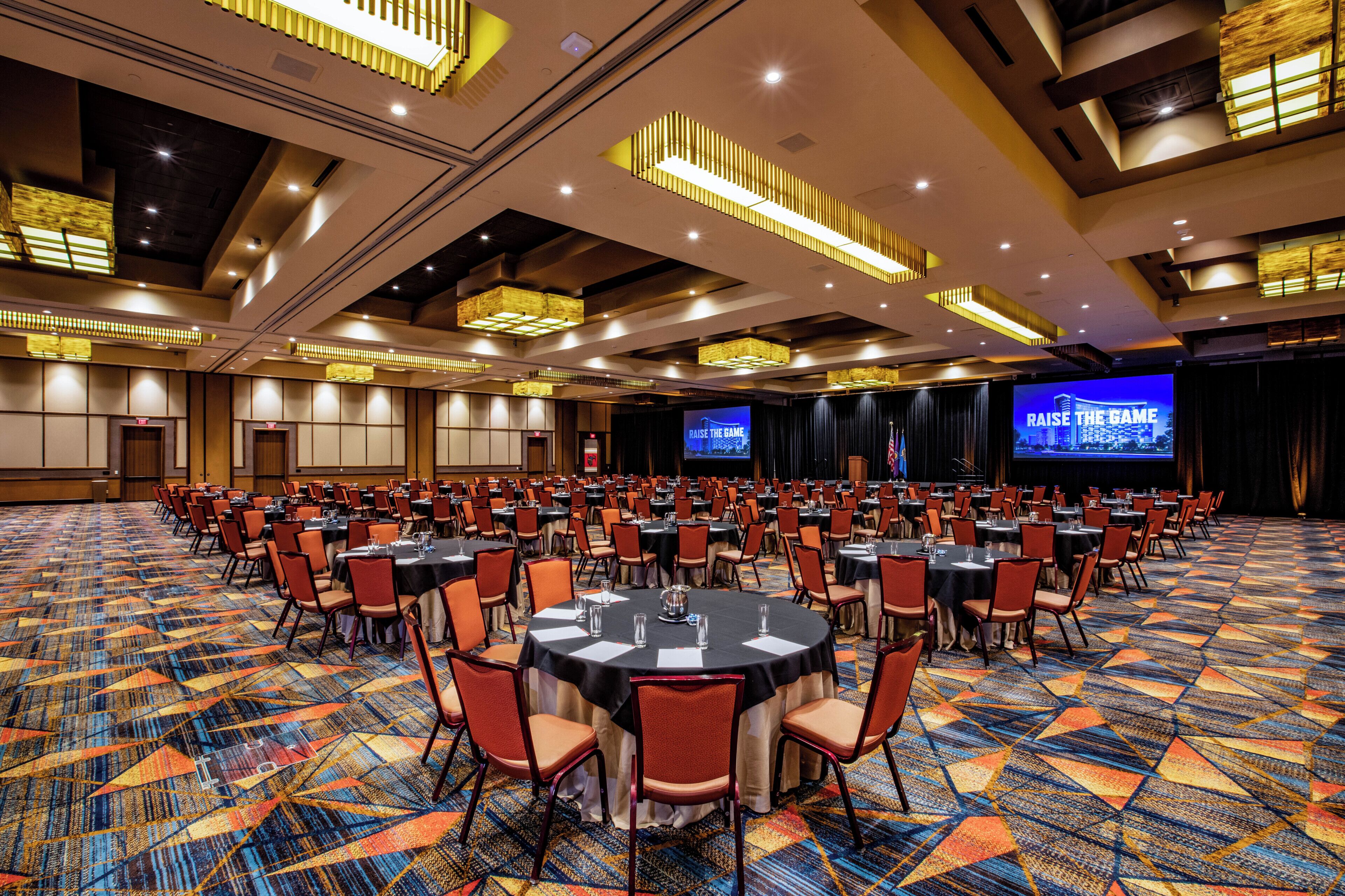 Un'ampia sala conferenze con tavoli rotondi, sedie e un palco con uno schermo che mostra 'RAISE THE GAME'.
