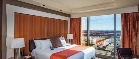 A hotel room with a large bed, two bedside tables, a desk with a chair, and a large window offering a city view.