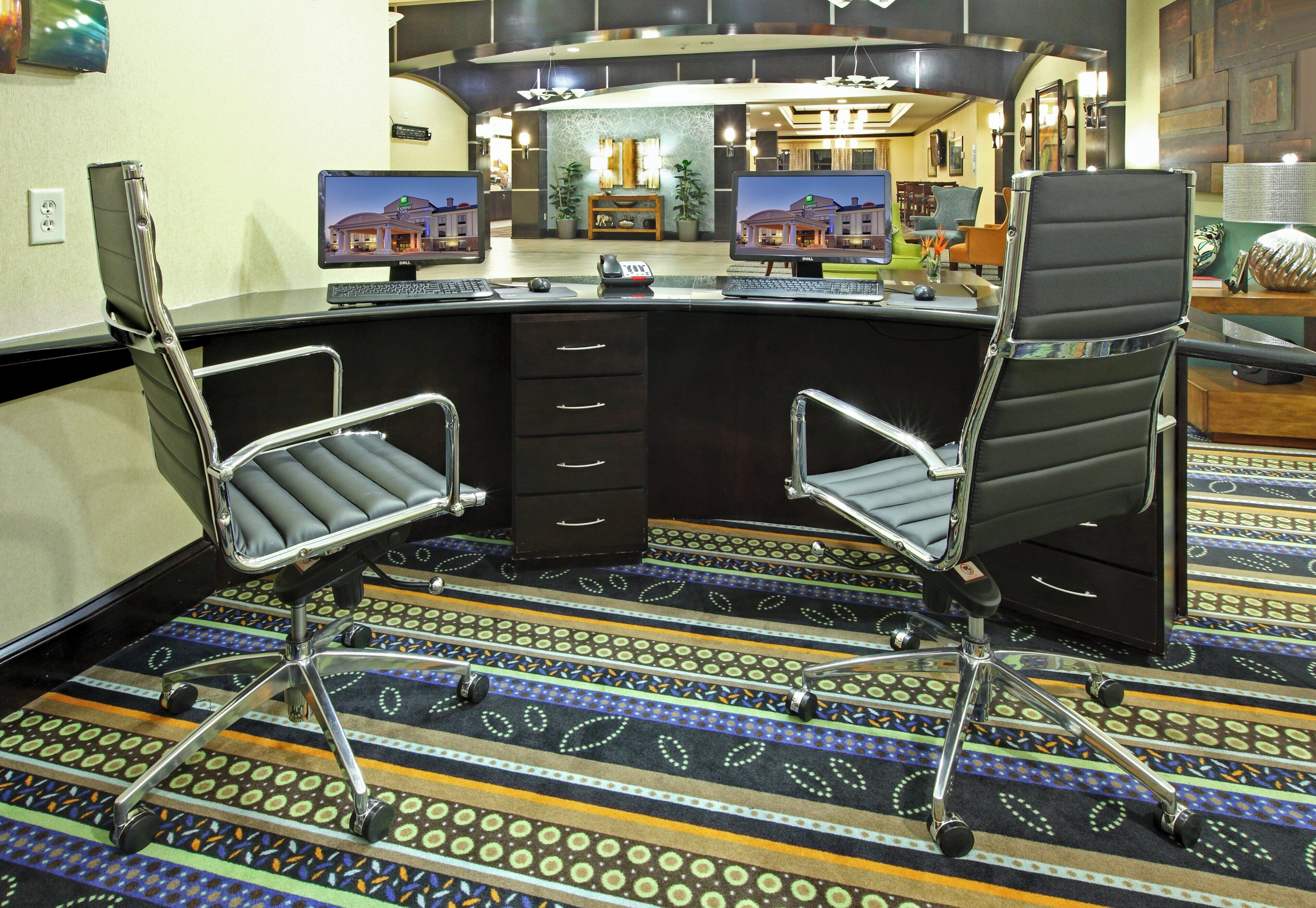 A hotel lobby with a reception desk, two office chairs, a patterned carpet, and a large mirror.