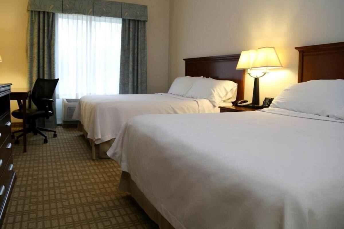 Habitación de hotel con dos camas, un escritorio, una silla, una lámpara y un ventanal con cortinas.