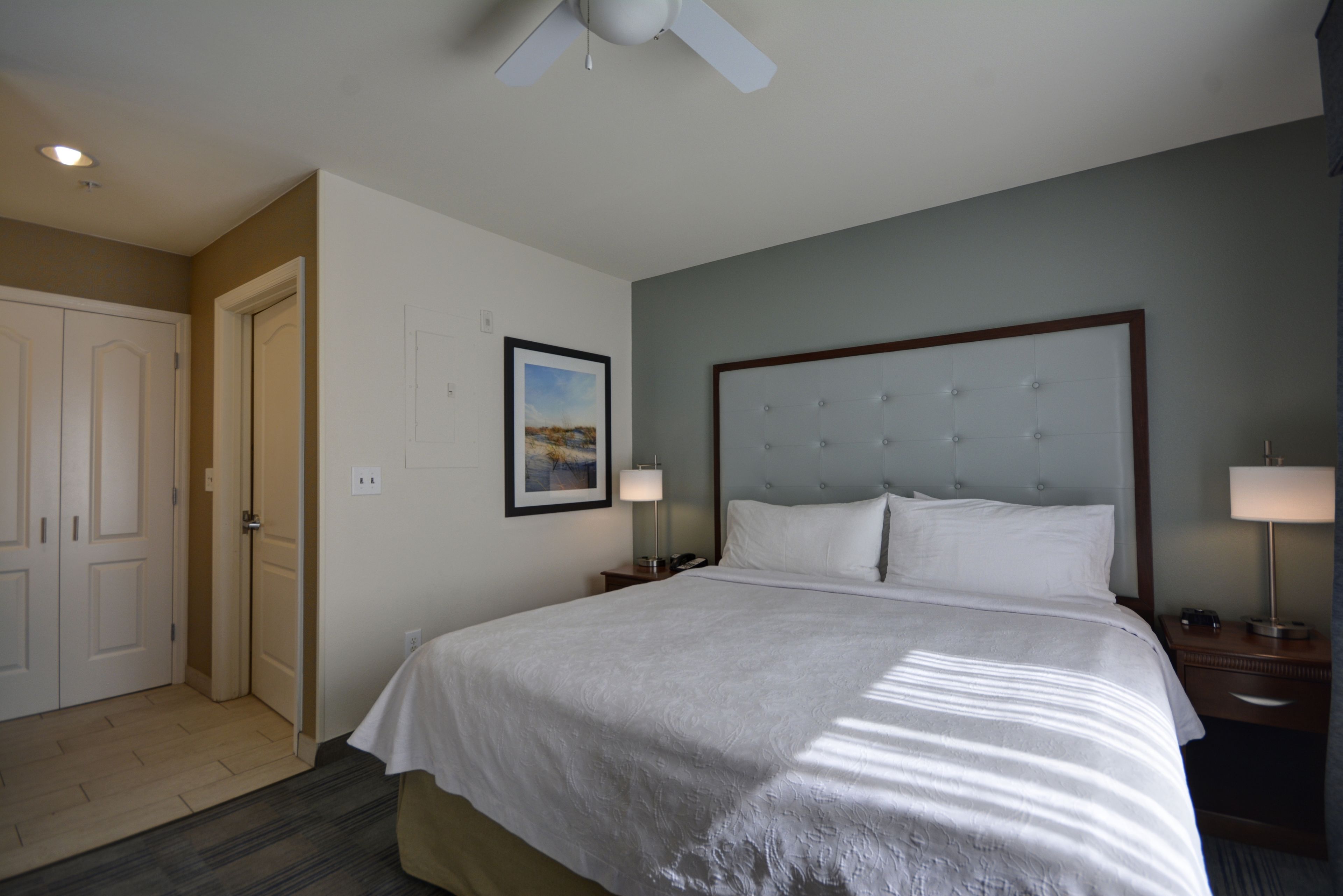 A hotel room with a large bed, a ceiling fan, a framed picture on the wall, and a door to the left.