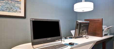 Una habitación de hotel con un escritorio, una computadora de escritorio, una impresora y un cuadro en la pared.