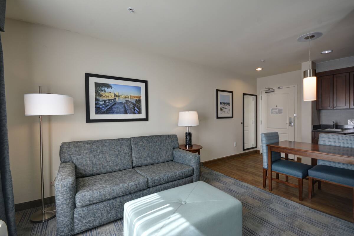 Una habitación de hotel moderna con un sofá gris, un pequeño taburete, una mesa de comedor con sillas y una zona de cocina.