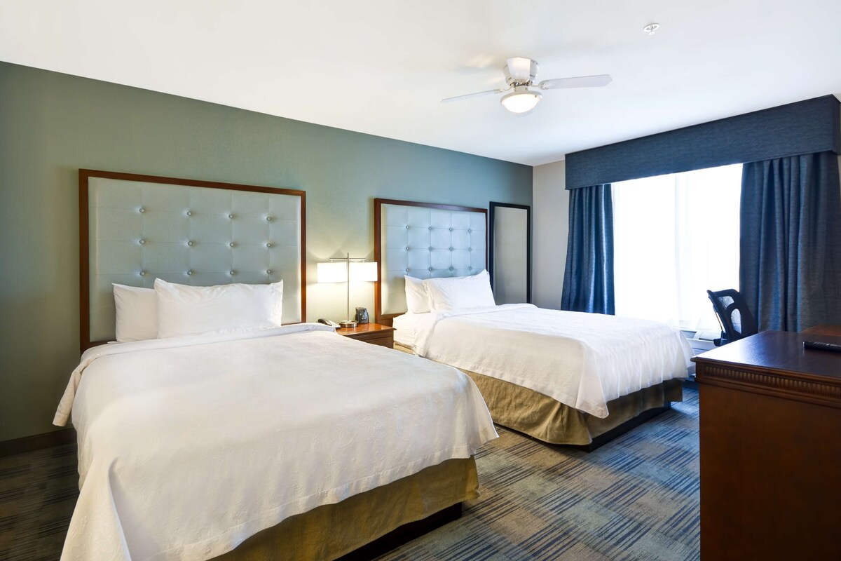 Une chambre d’hôtel avec deux lits, un bureau, une chaise, un ventilateur de plafond et une fenêtre avec des rideaux.