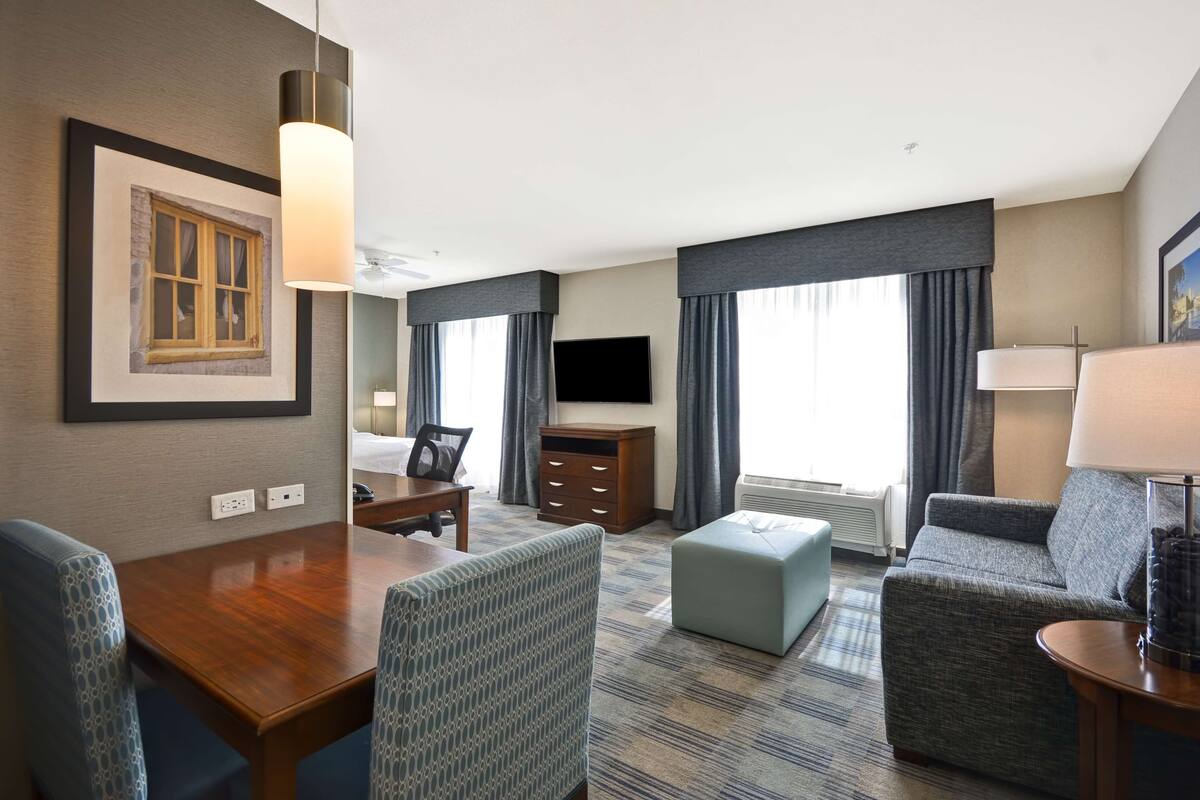 Habitación de hotel con una mesa de comedor de madera, dos sillas con estampado, un sofá, un televisor, un aparador y una ventana con cortinas.