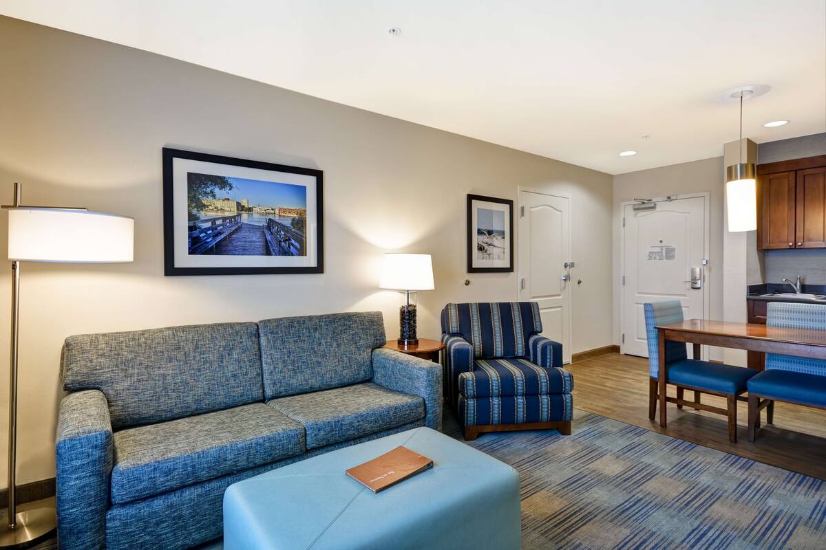 Una habitación de hotel moderna con un sofá gris, un sillón a rayas, una mesa de comedor con sillas azules y un cuadro enmarcado en la pared.