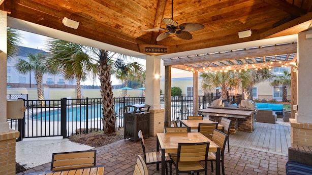 An outdoor patio with wooden tables and chairs, a ceiling fan, and a view of a swimming pool and palm trees. - Homewood Suites Wilmington/Mayfaire (Wilmington)