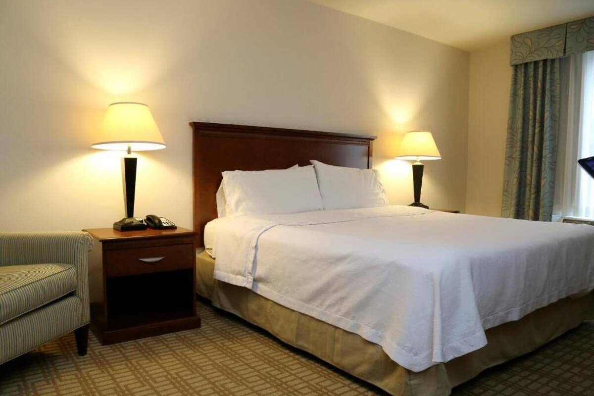 Habitación de hotel con cama, dos lámparas de noche, mesita de noche, silla y ventana con cortinas.