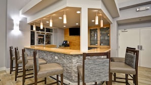 A modern bar area with a stone countertop, wooden chairs, pendant lighting, and a television. - Homewood Suites Wilmington/Mayfaire (Wilmington)