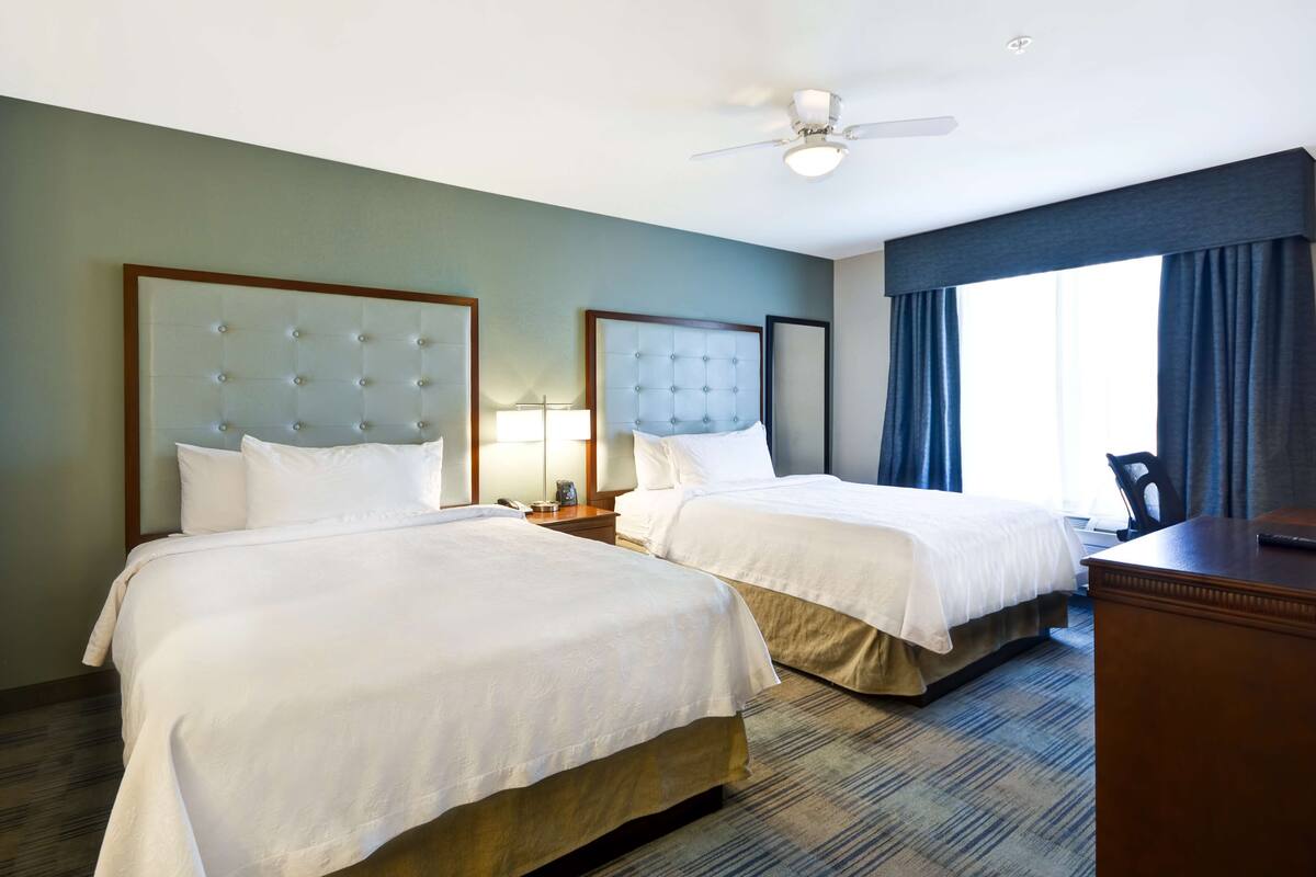 Habitación de hotel con dos camas, un escritorio, una silla, un ventilador de techo y una ventana con cortinas.