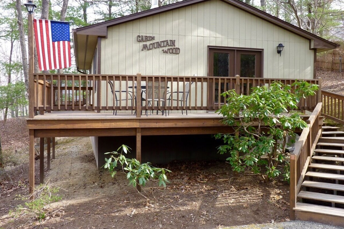 A cabin named Cabin Mountain Wood with a wooden deck, a flag, and a staircase leading to the entrance.