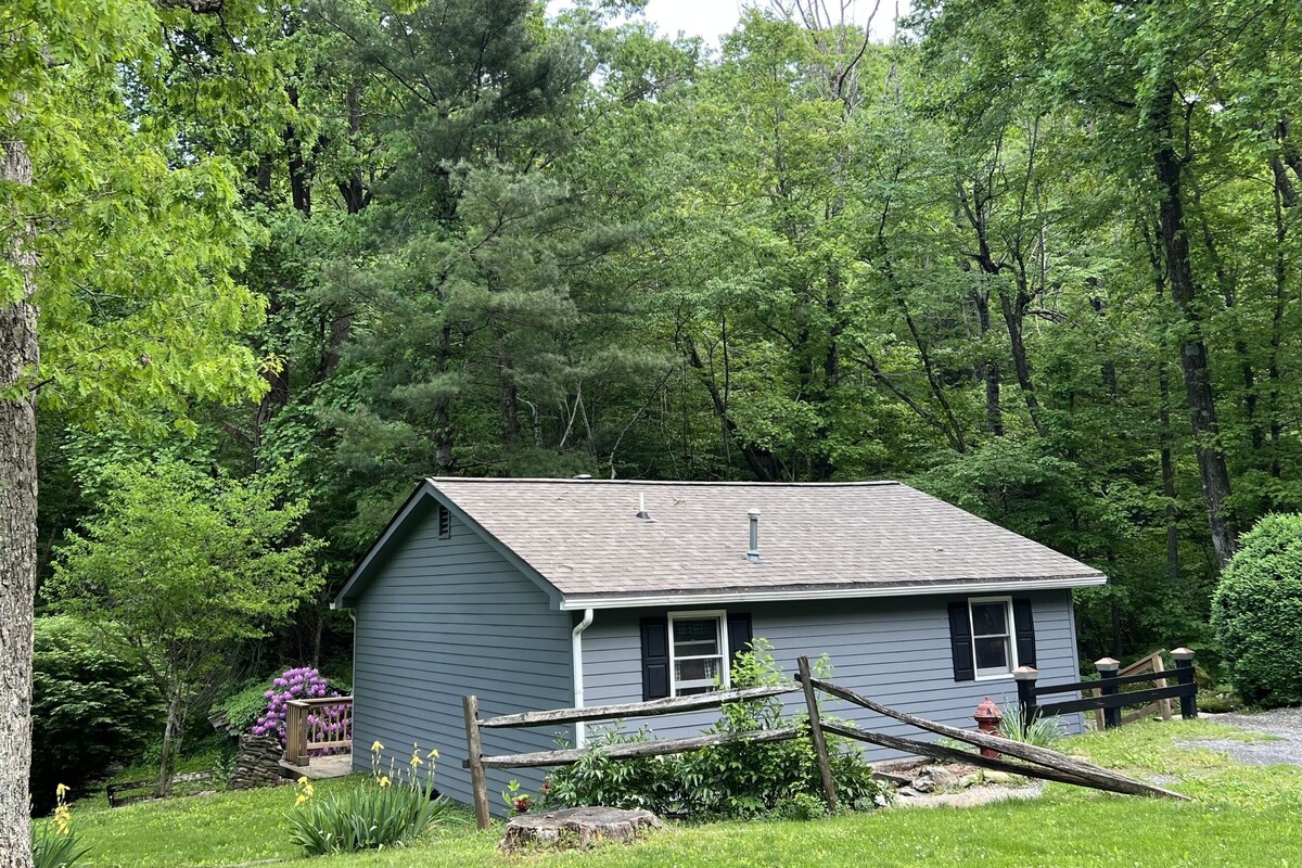 A small, single-story house with a sloped roof, surrounded by dense green trees and a well-maintained lawn.