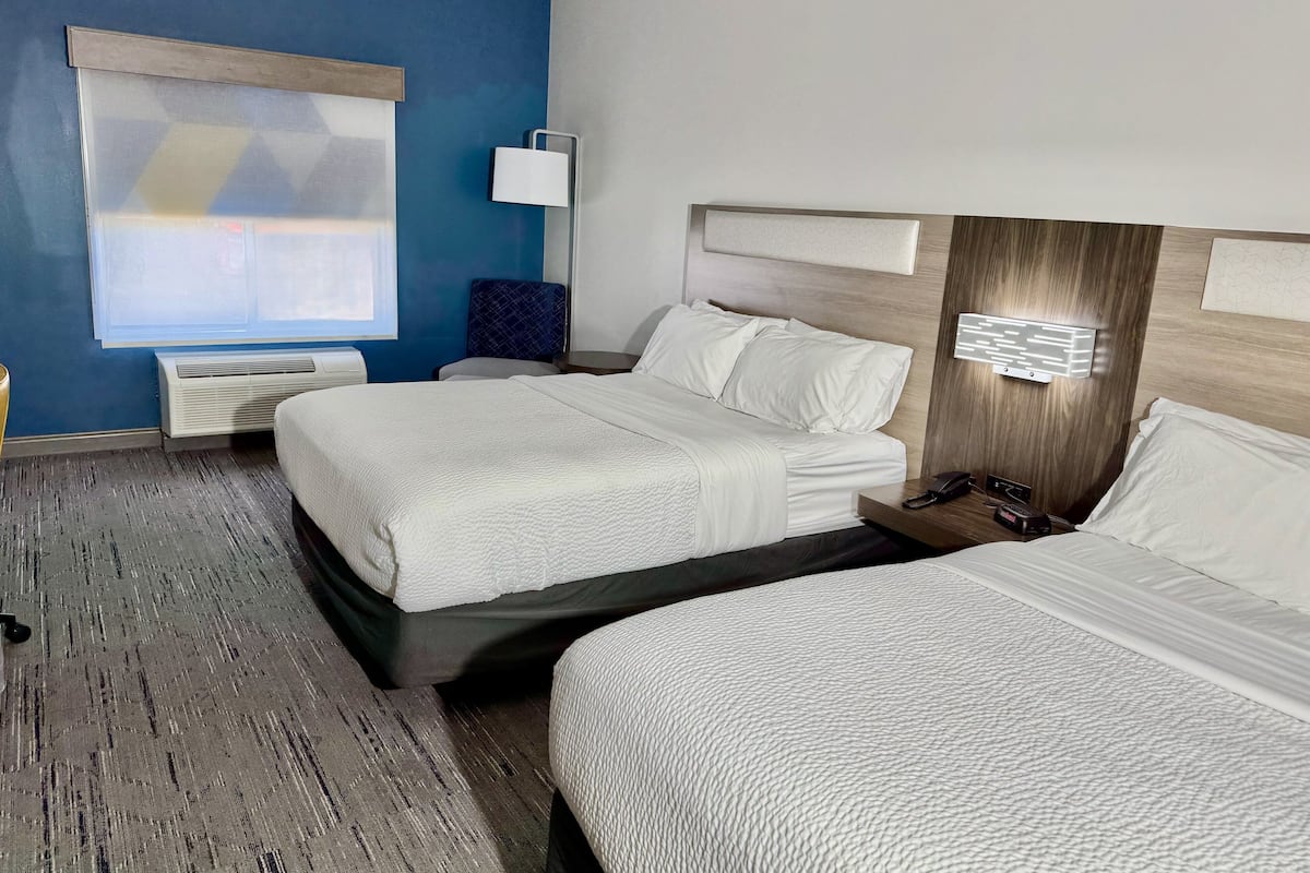 Habitación de hotel con dos camas, un escritorio, una silla, un ventanal con persianas y una lámpara de pared.
